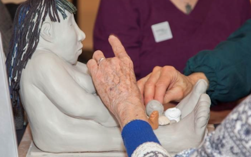 Close-up of an elderly person's hands engaging with a tactile sculpture of a seated figure with long, textured hair. The figure's outstretched legs form a bowl-like space, where small objects are placed. Other participants' hands are also interacting with the artwork in the background.